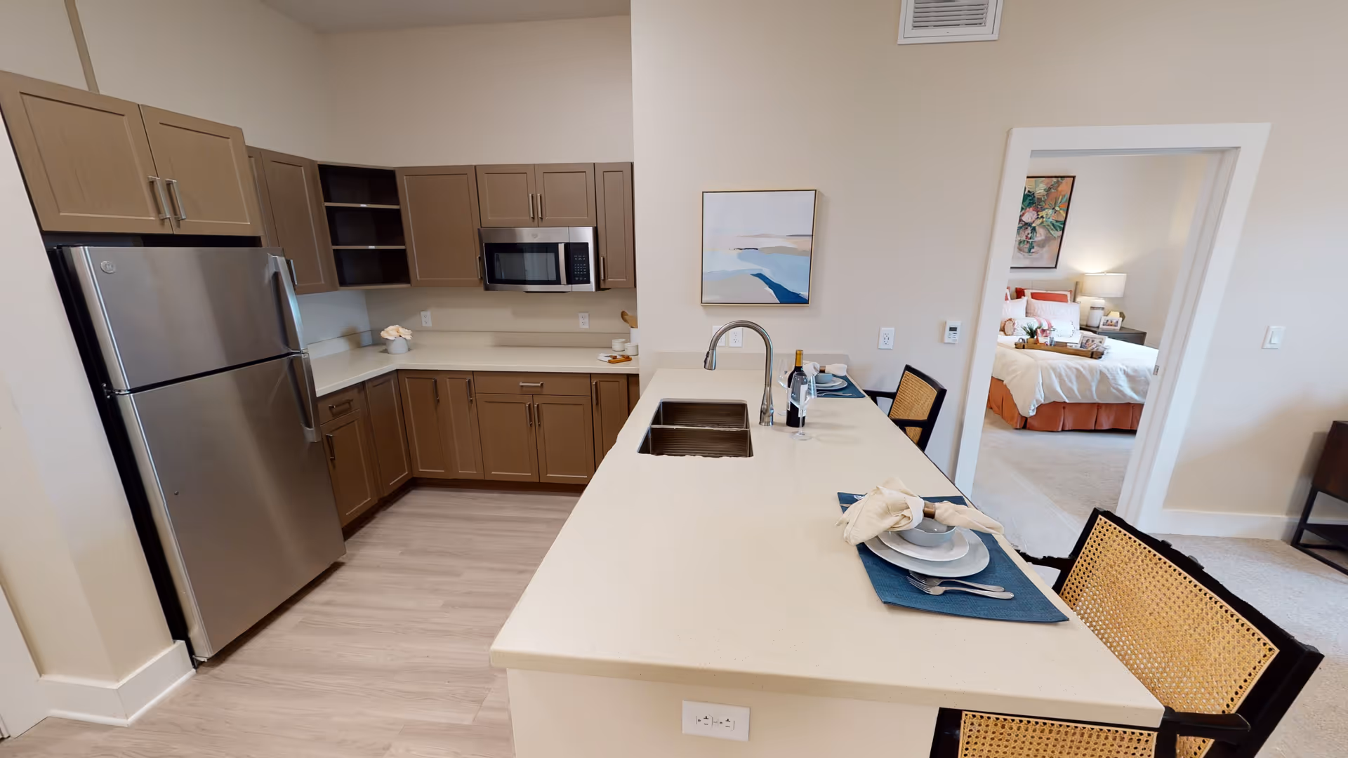 Modern kitchen with light brown cabinets, stainless steel refrigerator and microwave, a white countertop island with a sink, two place settings with plates, bowls, napkins, and utensils, and a bottle of wine with two glasses. In the background, a doorway leads to a bedroom with a bed, nightstand, lamp, and artwork on the wall.