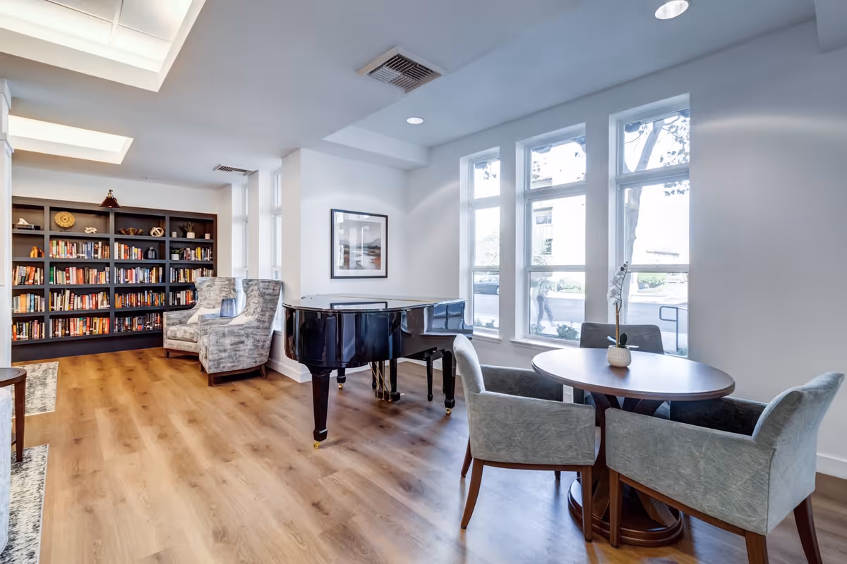 A bright and spacious room with large windows letting in natural light. The room features a black grand piano in the center, a round wooden table with four upholstered chairs, and a bookshelf filled with books against the far wall. Two patterned armchairs are placed near the bookshelf, and a framed picture hangs on the wall above the piano. The floor is wooden, and the ceiling has recessed lighting.