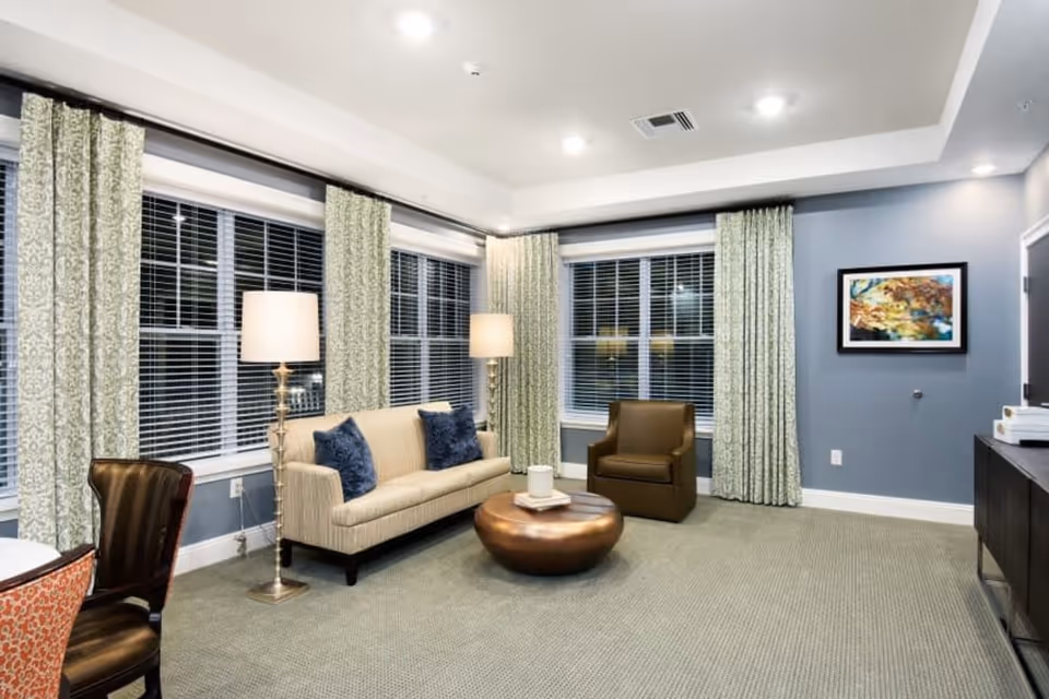 Well-lit communal living room with a sofa, armchair, two floor lamps, round coffee table, large windows with patterned curtains, and framed artwork on the wall.