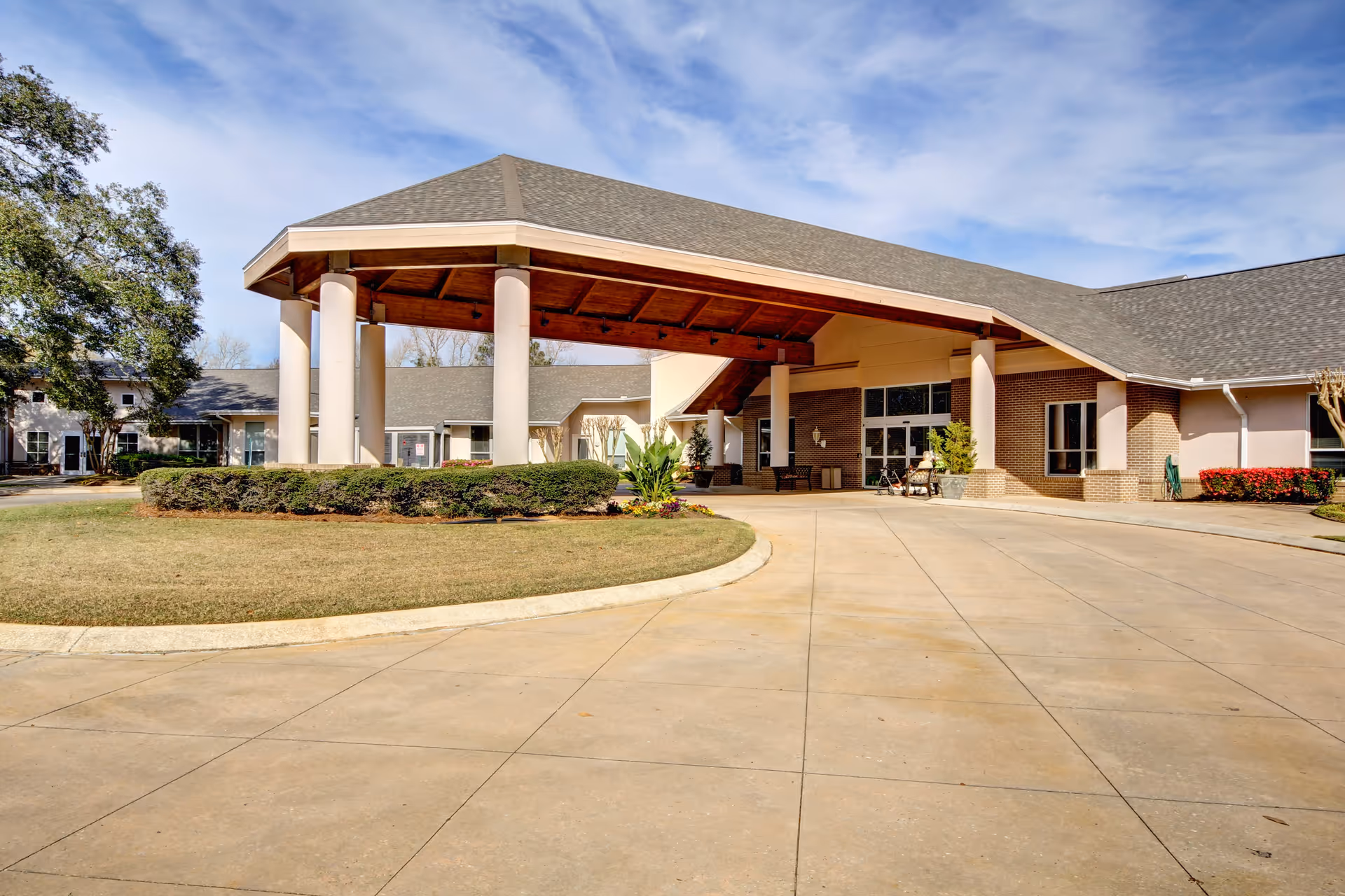 Entrance canopy and driveway of a single-story senior living facility building with columns and landscaping under a blue sky.