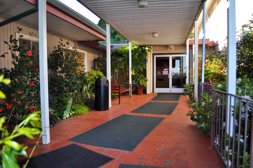 Covered entrance walkway with mats, benches, plants and double glass doors leading into the senior living building.