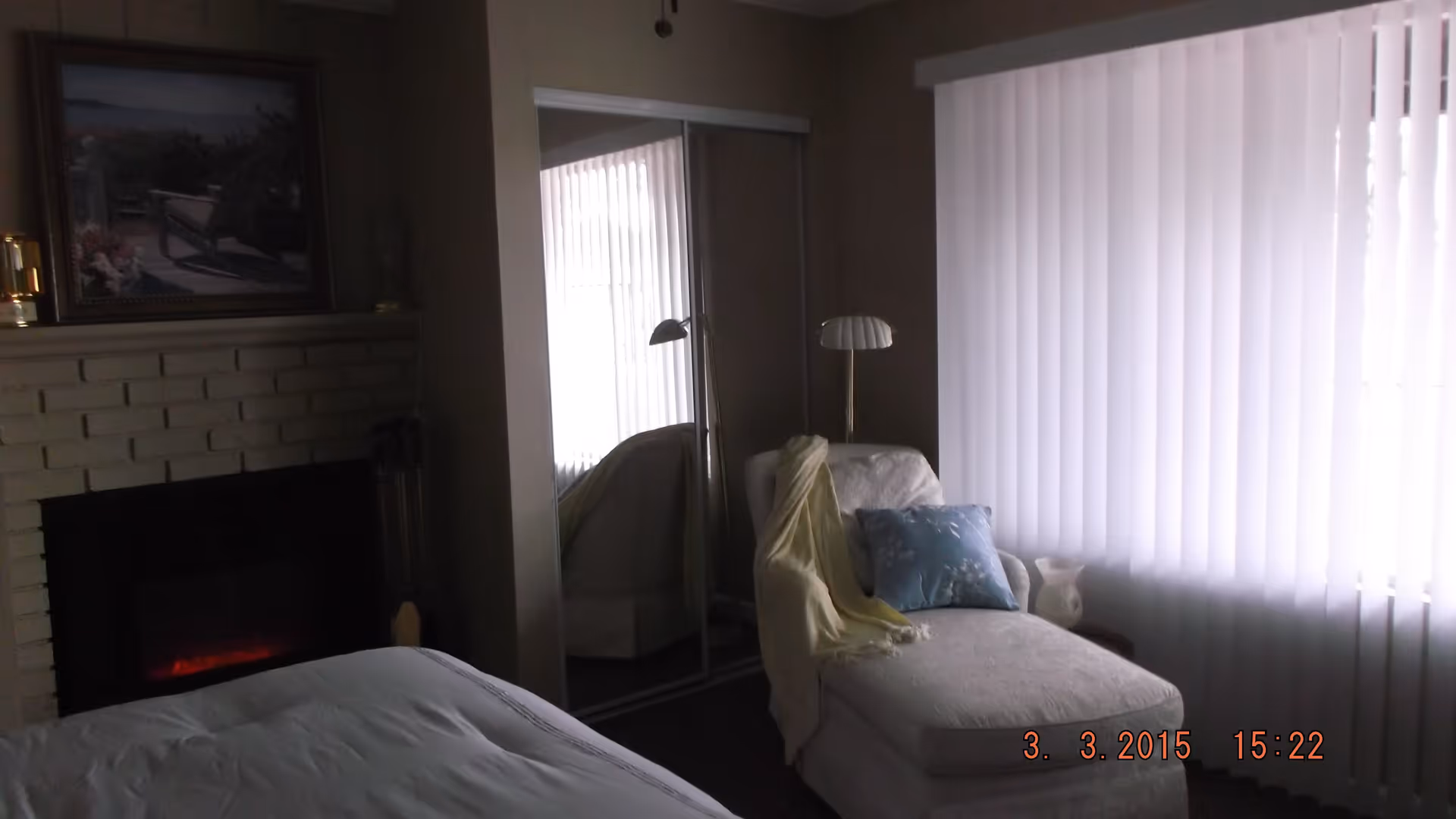 A cozy bedroom featuring a bed with white bedding, a white brick fireplace with a painting above it, a white chaise lounge with a blue pillow and a yellow throw blanket, a floor lamp, and vertical blinds covering a large window.