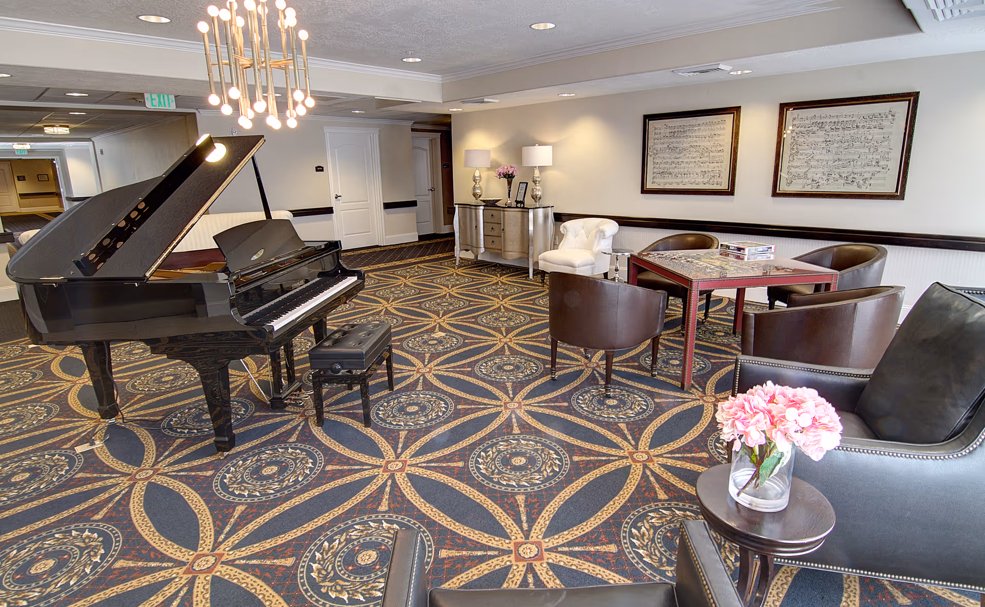 A spacious common area with a grand piano and piano bench on a patterned carpet. The room features a seating area with a table and four chairs, two framed musical score artworks on the wall, a white armchair, a sideboard with two lamps and flowers, and a small round table with a vase of pink flowers in the foreground.