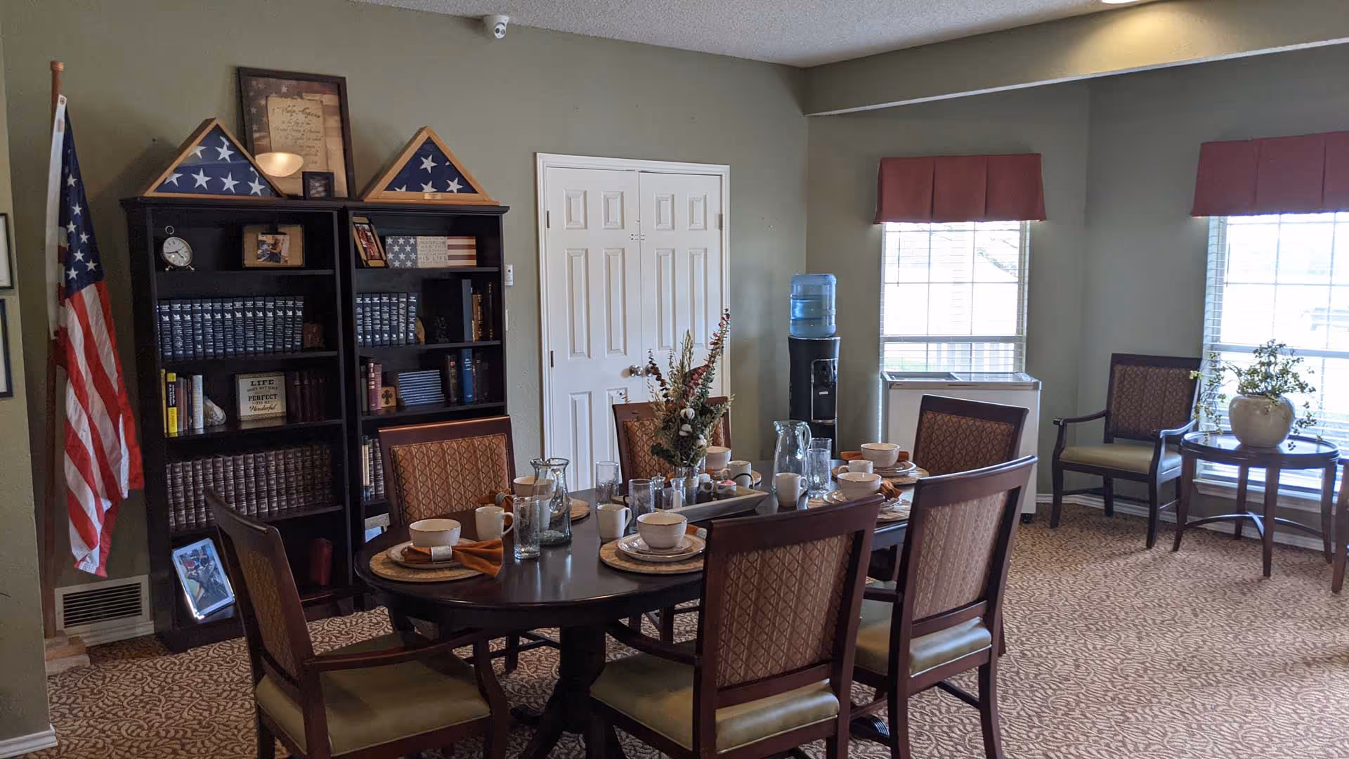 A dining room with a dark wooden table set for six people with bowls, plates, glasses, and napkins. There are six upholstered chairs around the table. In the background, there is a black bookshelf filled with books, framed pictures, and two folded American flags in triangular display cases. An American flag stands to the left of the bookshelf. Two windows with red valances let in natural light, and a water cooler is positioned near the windows. A small round table with a plant and two chairs is near the windows. The walls are painted green, and the carpet has a patterned design.