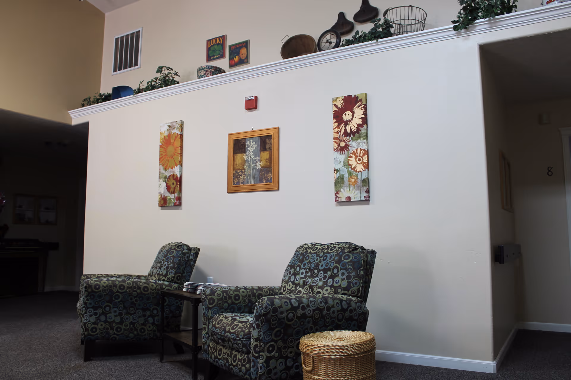 Two patterned armchairs and a small side table in a seating area with floral wall art and decorative items on a ledge above.