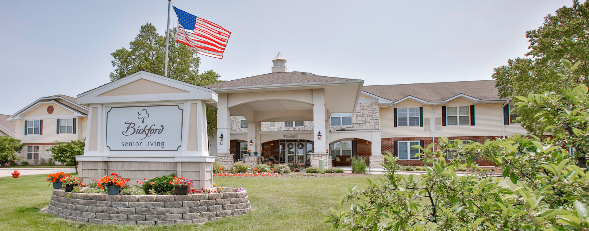 Front exterior of the Bickford senior living building with a covered entrance, landscaped lawn and an American flag.