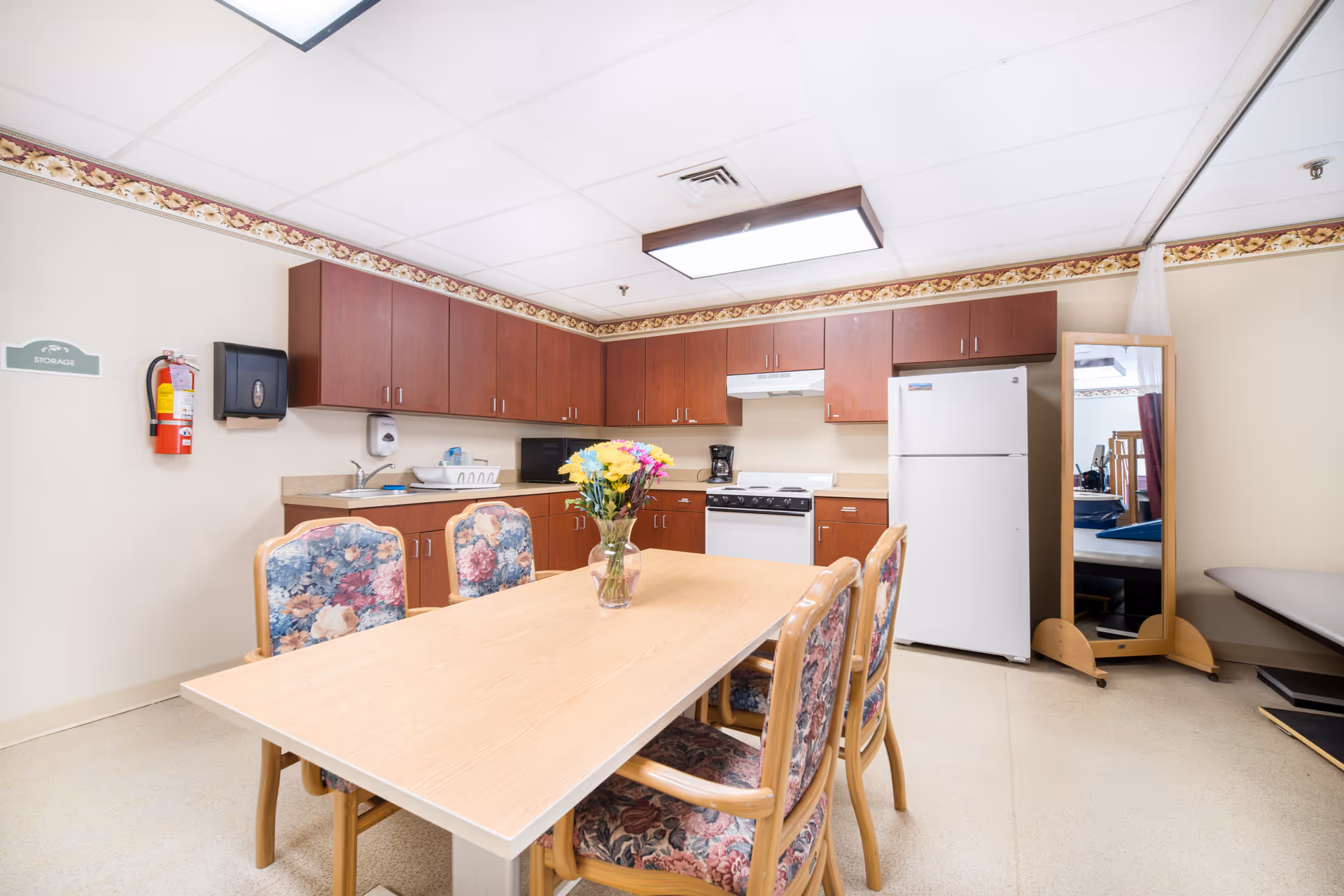 Communal dining area with a wooden table and floral-upholstered chairs in front of a kitchenette with cabinets, refrigerator, and stove.