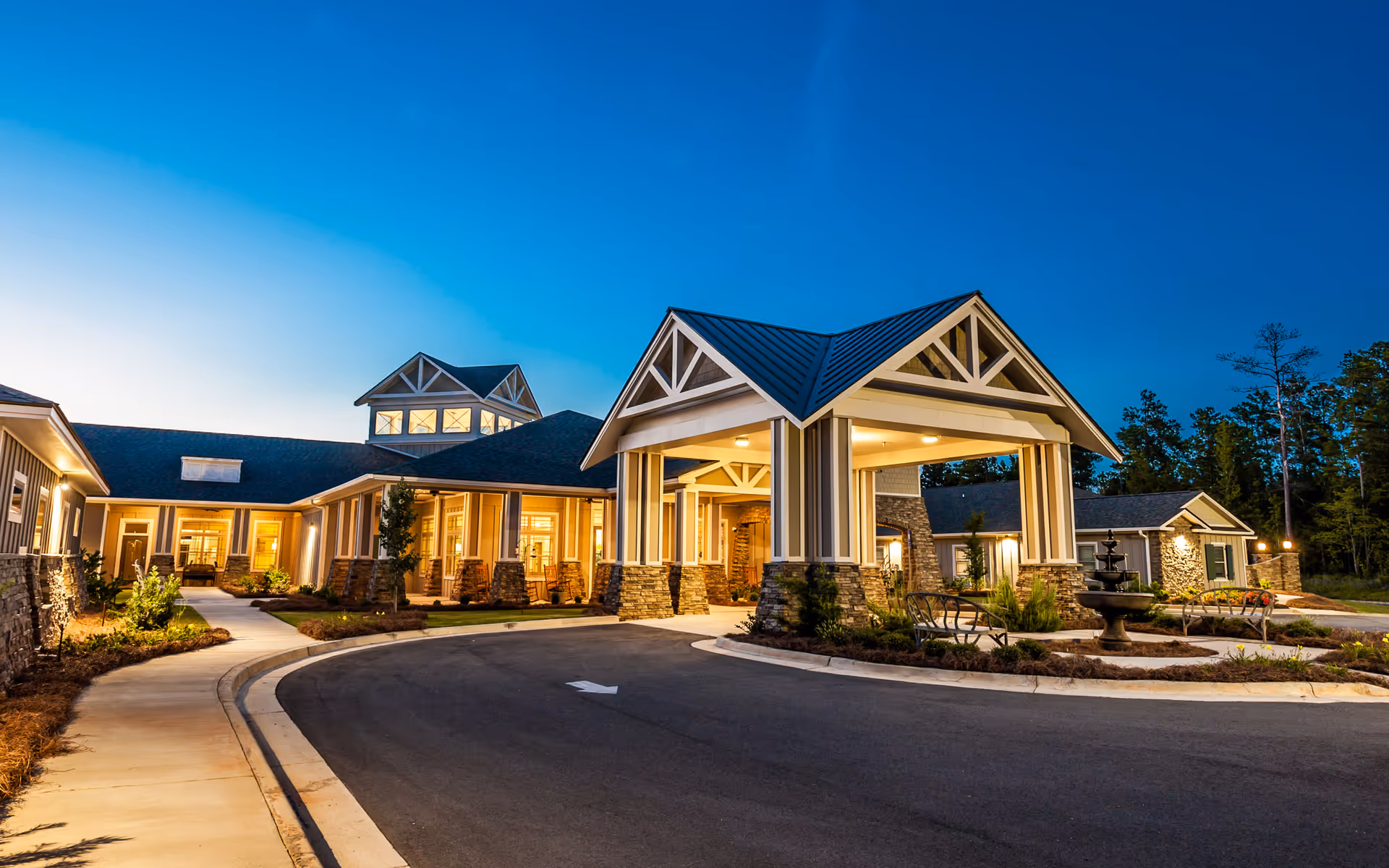 Exterior view of Fellowship Meriwether Assisted Living Community at dusk, showing a well-lit entrance with a covered driveway, stone and wood architectural details, landscaped surroundings, and a fountain near the entrance.