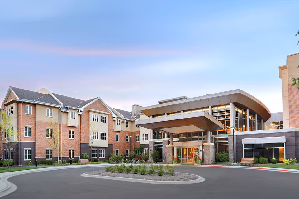 Exterior view of Hearthstone of Arlington Heights senior living facility showing a modern multi-story building with brick and beige siding, large windows, and a covered entrance with stone pillars. There is a circular driveway with landscaping in front of the entrance under a clear sky at dusk.