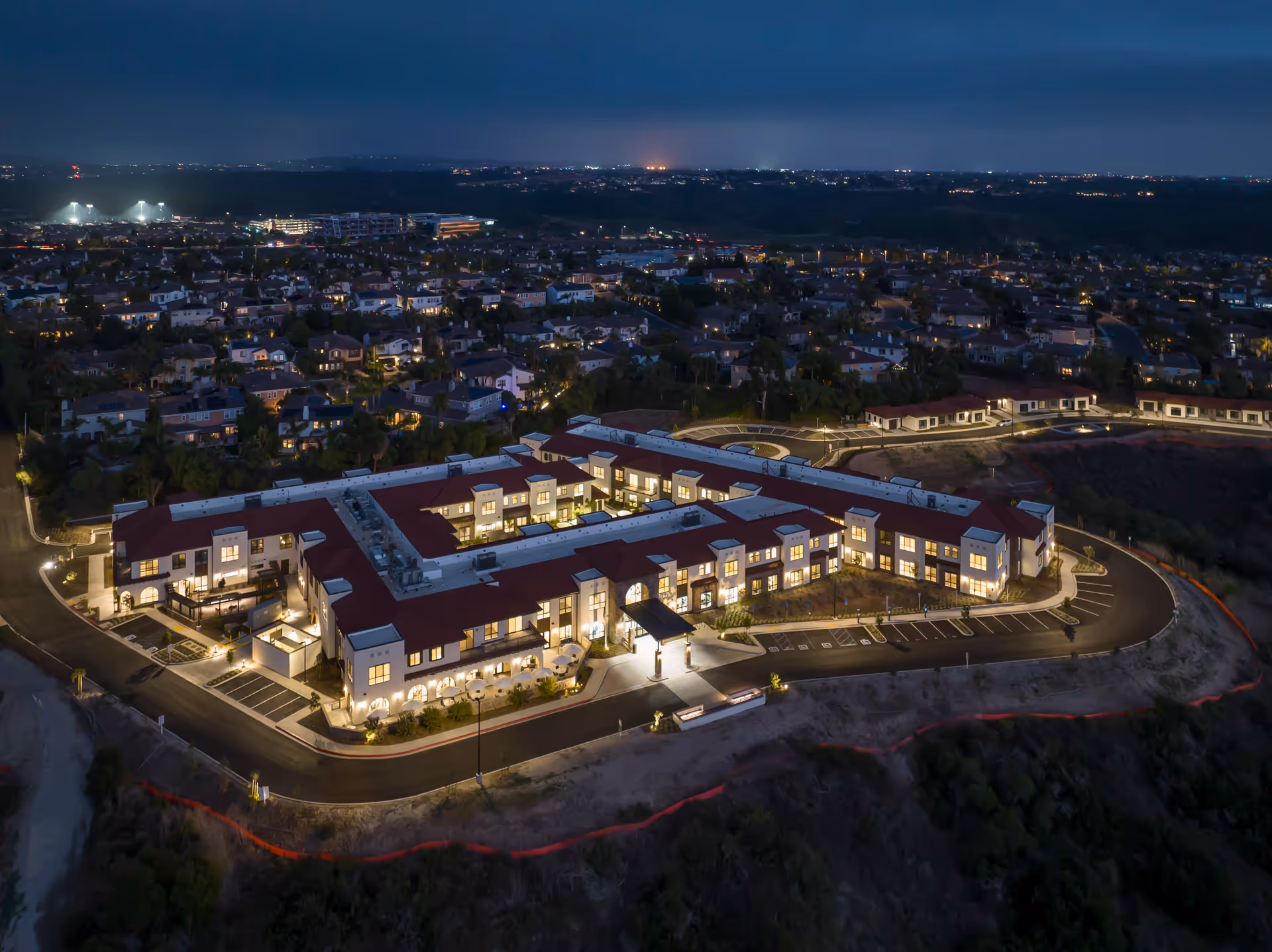 Aerial nighttime view of Westmont of Carmel Valley, a large illuminated senior living facility with a red roof, surrounded by a residential neighborhood and hills in the background.