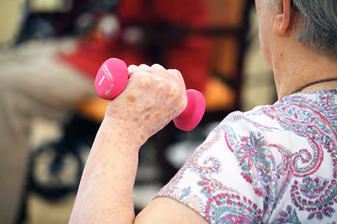 An elderly person lifting a pink 1-pound dumbbell during an exercise session, wearing a patterned shirt with a paisley design.