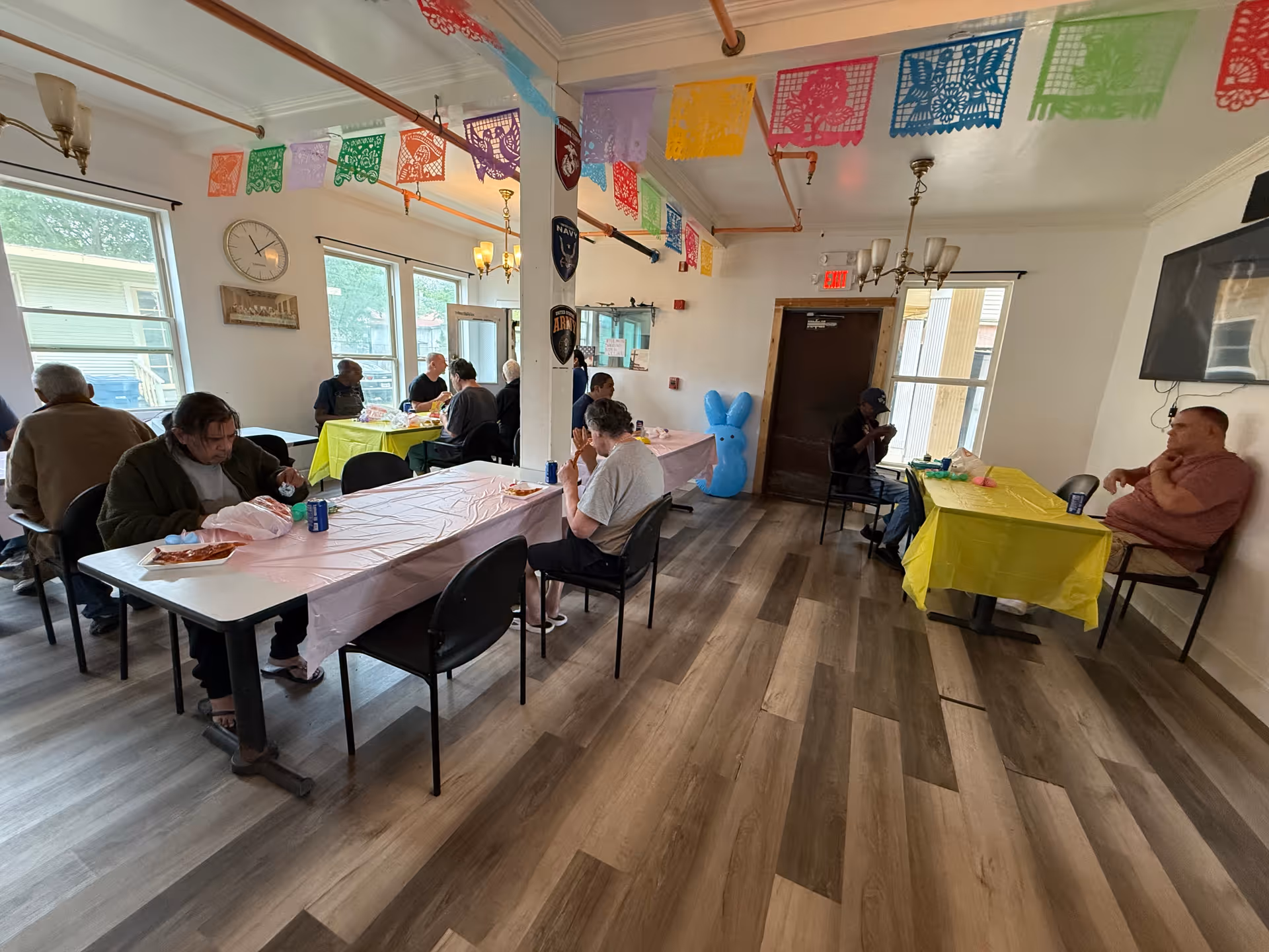 Several people seated at long tables covered with tablecloths in a decorated communal dining/activity room.