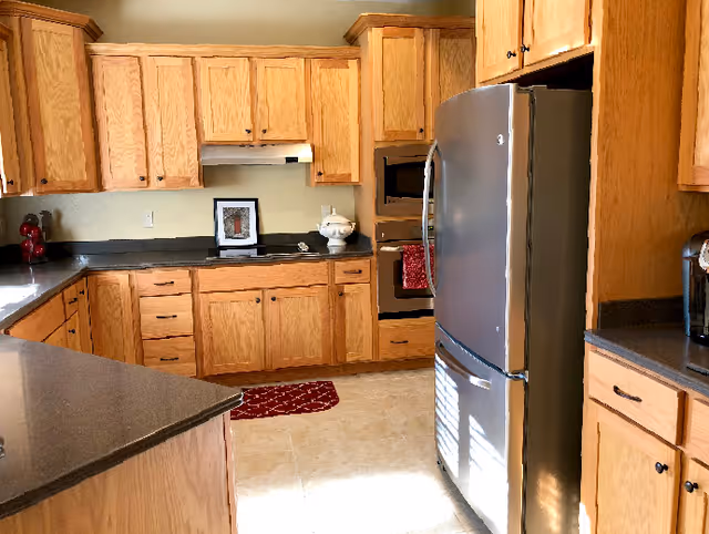 A kitchen with wooden cabinets and drawers, a stainless steel refrigerator, a built-in oven, a microwave, and dark countertops. There is a red rug on the tiled floor and a few kitchen items on the counters including a red mixer and a white decorative bowl.