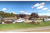 Exterior view of Rolling Hills Healthcare and Rehabilitation Center, a single-story building with a brown roof and beige walls. Several cars are parked in front of the building along the driveway. An American flag is flying on a flagpole near the entrance, with a sign below it. The sky is blue with some clouds, and there is green grass and trees around the building.