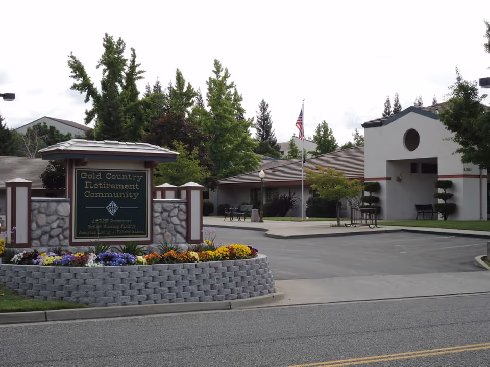 Entrance to Gold Country Retirement Community showing the main sign, a flower bed, flagpole, and the building driveway.