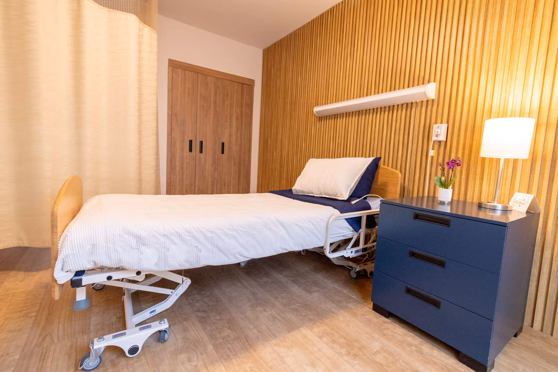 A simple patient bedroom with a hospital bed, blue three-drawer nightstand holding a lamp and small plant, wood-paneled wall, and a curtained closet.