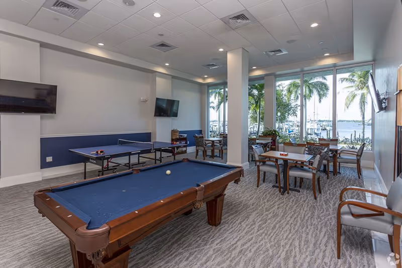A recreational room with a pool table and a ping pong table, several chairs and tables, large windows showing palm trees and a waterfront view outside.