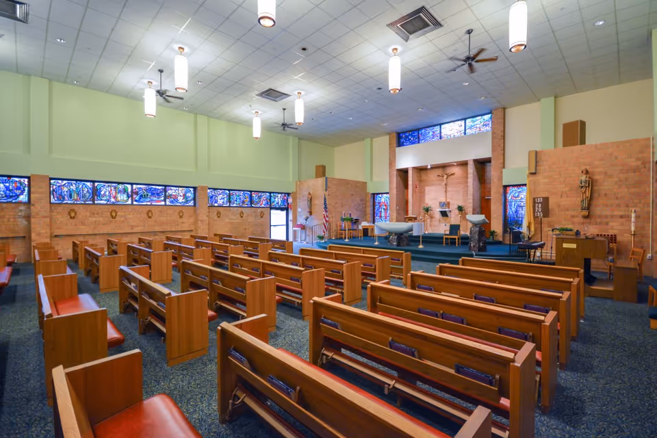 Interior view of a chapel with wooden pews arranged in rows facing an altar. The altar area has religious statues, a crucifix on the wall, and stained glass windows above and along the side walls. The ceiling has hanging lights and ceiling fans.