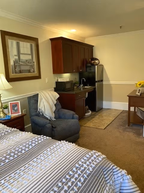 A cozy assisted living room featuring a bed with a striped and textured blanket, a comfortable blue recliner chair with a white throw draped over it, a wooden nightstand with a lamp and framed photo, a kitchenette area with dark wood cabinets, a microwave, a sink, and a stainless steel refrigerator. There is a framed picture on the wall and a wooden table with a vase of sunflowers and a picture frame on the right side.