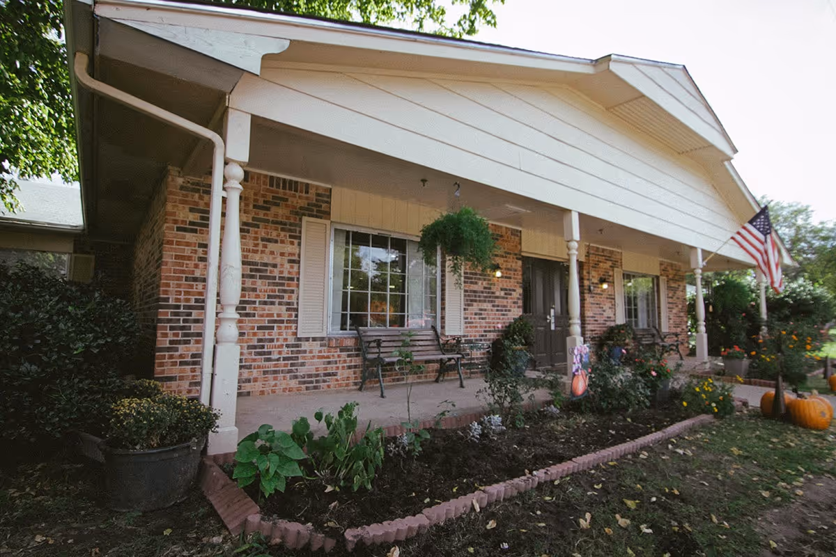 Front exterior view of a brick nursing center building with a covered porch, benches, hanging plants, garden beds, and an American flag displayed on the right side.