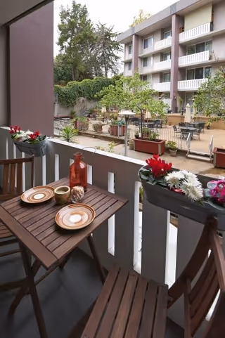 A small balcony with a wooden bistro table set for two overlooking a landscaped courtyard and multi-story senior living building.