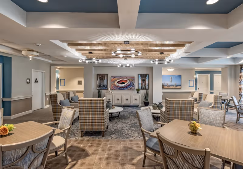 A spacious and well-lit common area in a senior living facility featuring multiple seating arrangements with plaid and cushioned chairs around small tables. The room has modern ceiling lights with wooden beams and decorative wall art including a stadium and architectural images. The walls are painted in neutral tones with blue accents, and there are doors leading to other rooms in the background.