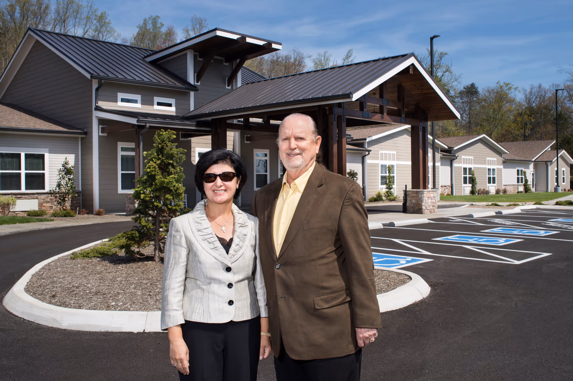 A man and a woman standing outside in front of a modern assisted living facility building with a covered entrance and several handicap parking spaces visible. The woman is wearing sunglasses and a light-colored blazer, and the man is wearing a brown jacket and yellow shirt. Trees and a clear blue sky are in the background.