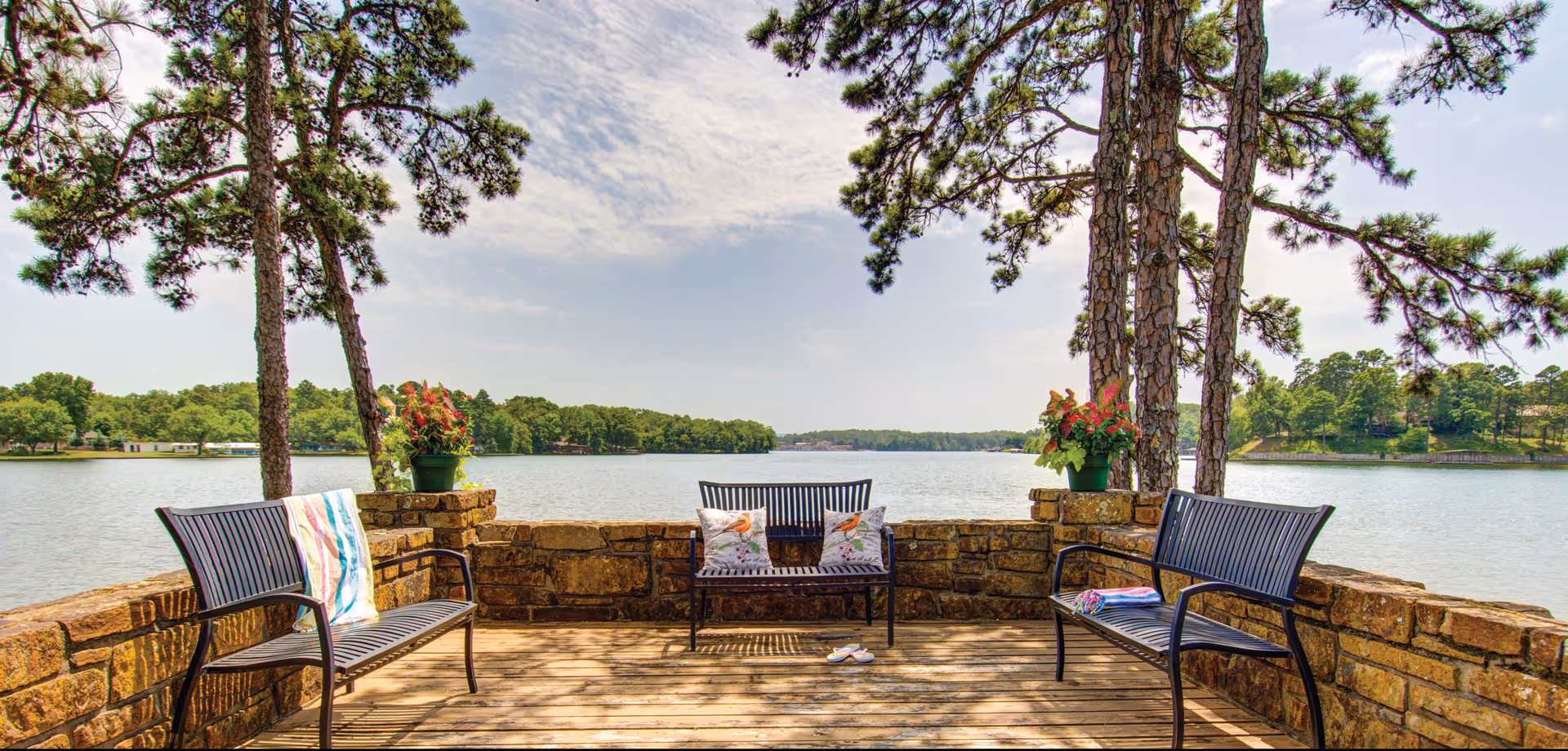 A peaceful outdoor seating area overlooking a large lake, featuring three black metal benches with cushions and a towel, surrounded by a low stone wall and tall pine trees under a partly cloudy sky.