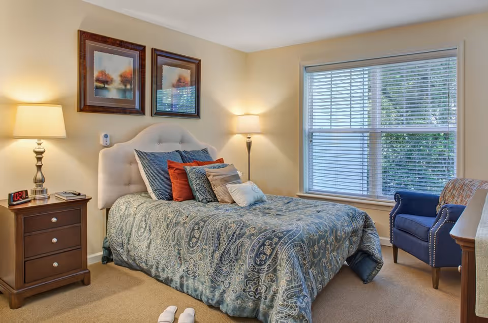 A cozy bedroom with a bed featuring a patterned blue and gray comforter and multiple pillows in blue, red, and beige. There are two framed pictures of autumn trees above the tufted headboard. On the left side of the bed is a wooden nightstand with a silver lamp, a digital clock showing 10:21, and a phone. On the right side is a tall floor lamp. A large window with white blinds lets in natural light, and a blue armchair with nailhead trim is positioned near the window. A pair of white slippers is placed on the carpeted floor near the foot of the bed.