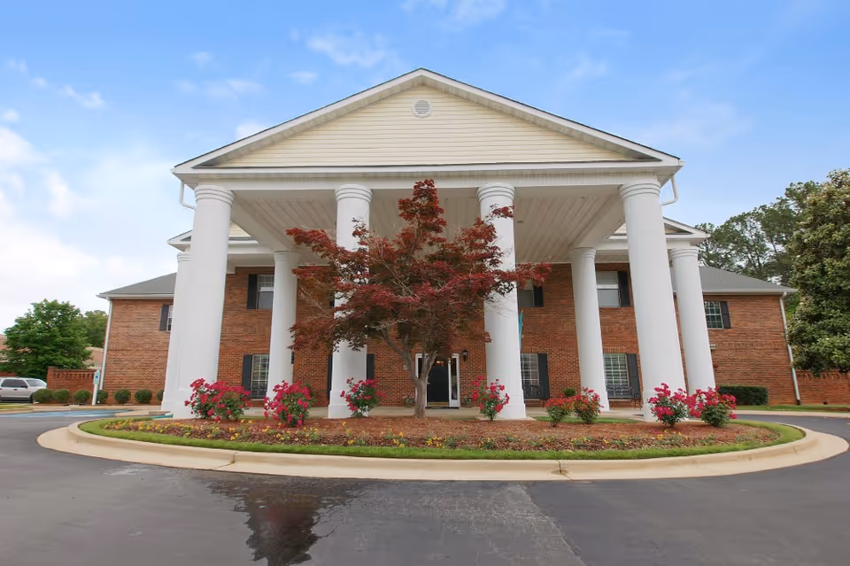 Front exterior view of a two-story brick building with large white columns supporting a covered entrance. There is a circular driveway with landscaped flower beds and a tree in front of the entrance. The sky is partly cloudy.