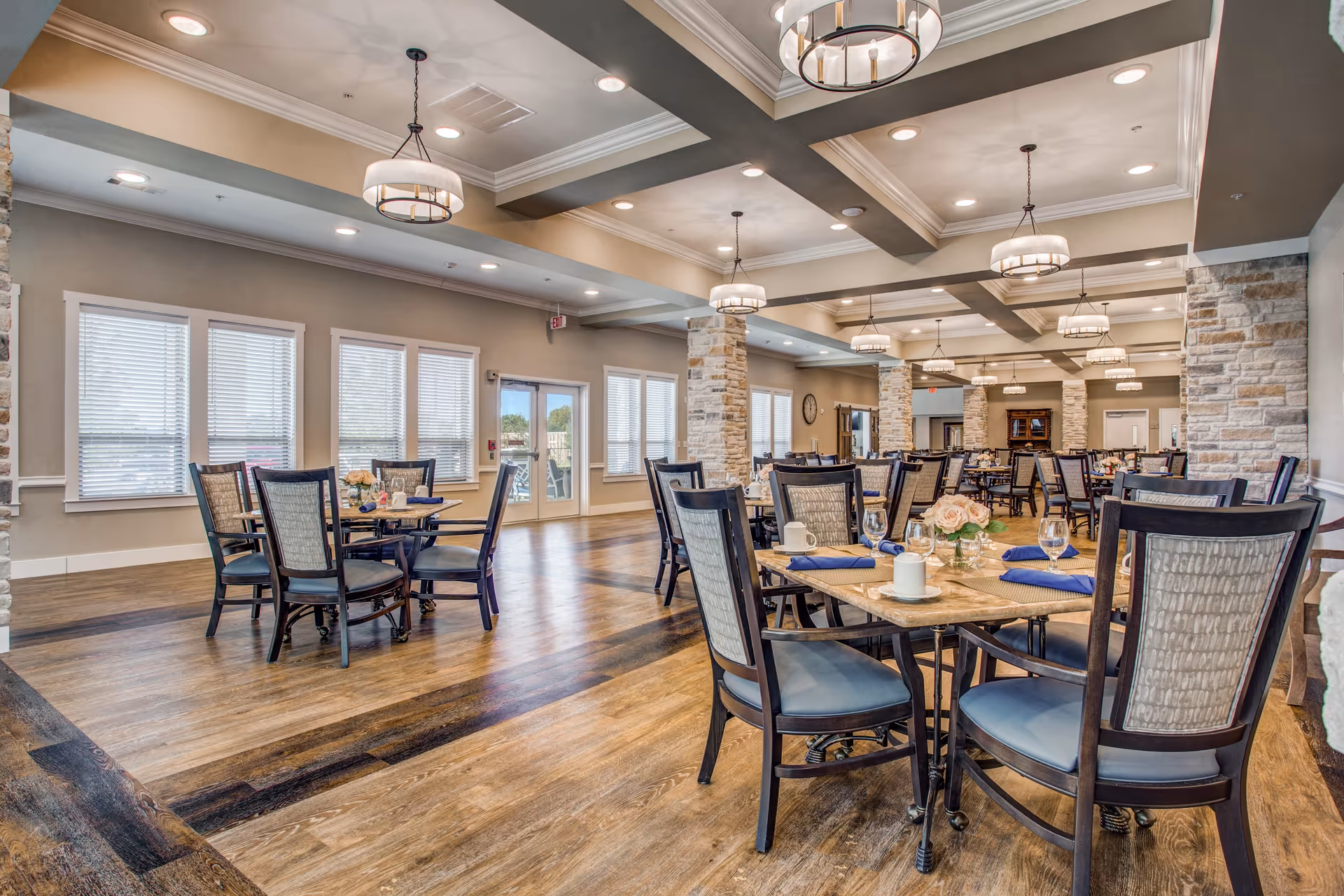 A spacious dining room with multiple tables and chairs arranged neatly. Each table is set with blue napkins, glassware, and floral centerpieces. The room features wooden flooring, stone pillars, large windows with blinds, and modern ceiling lights.