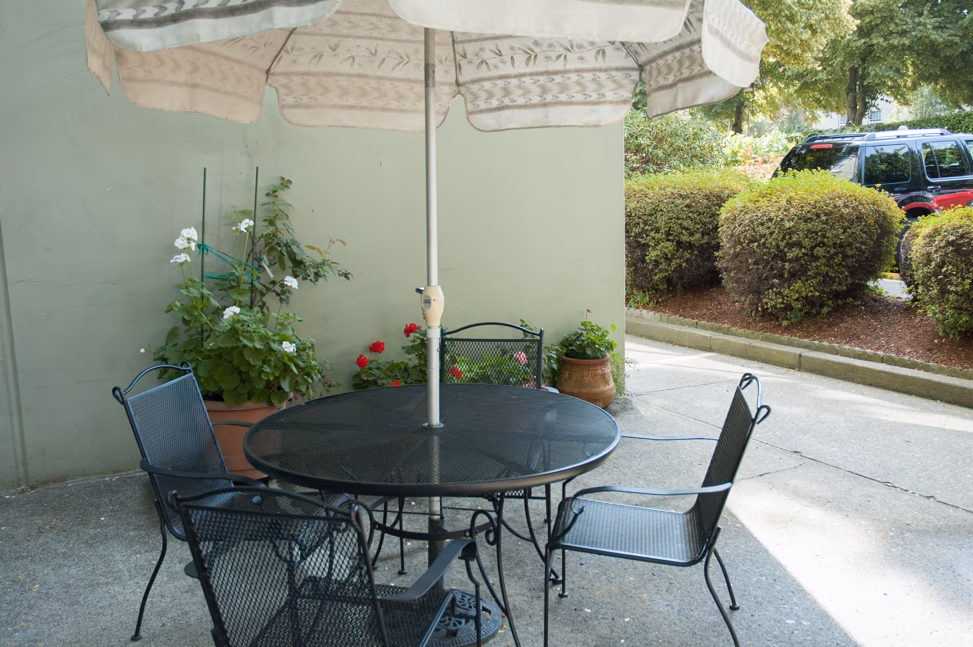 Outdoor patio area with a round black metal table and four matching chairs. A large beige umbrella is positioned in the center of the table. There are potted plants with green foliage and flowers against a light green wall. In the background, there are trimmed bushes and a black SUV parked on a driveway.