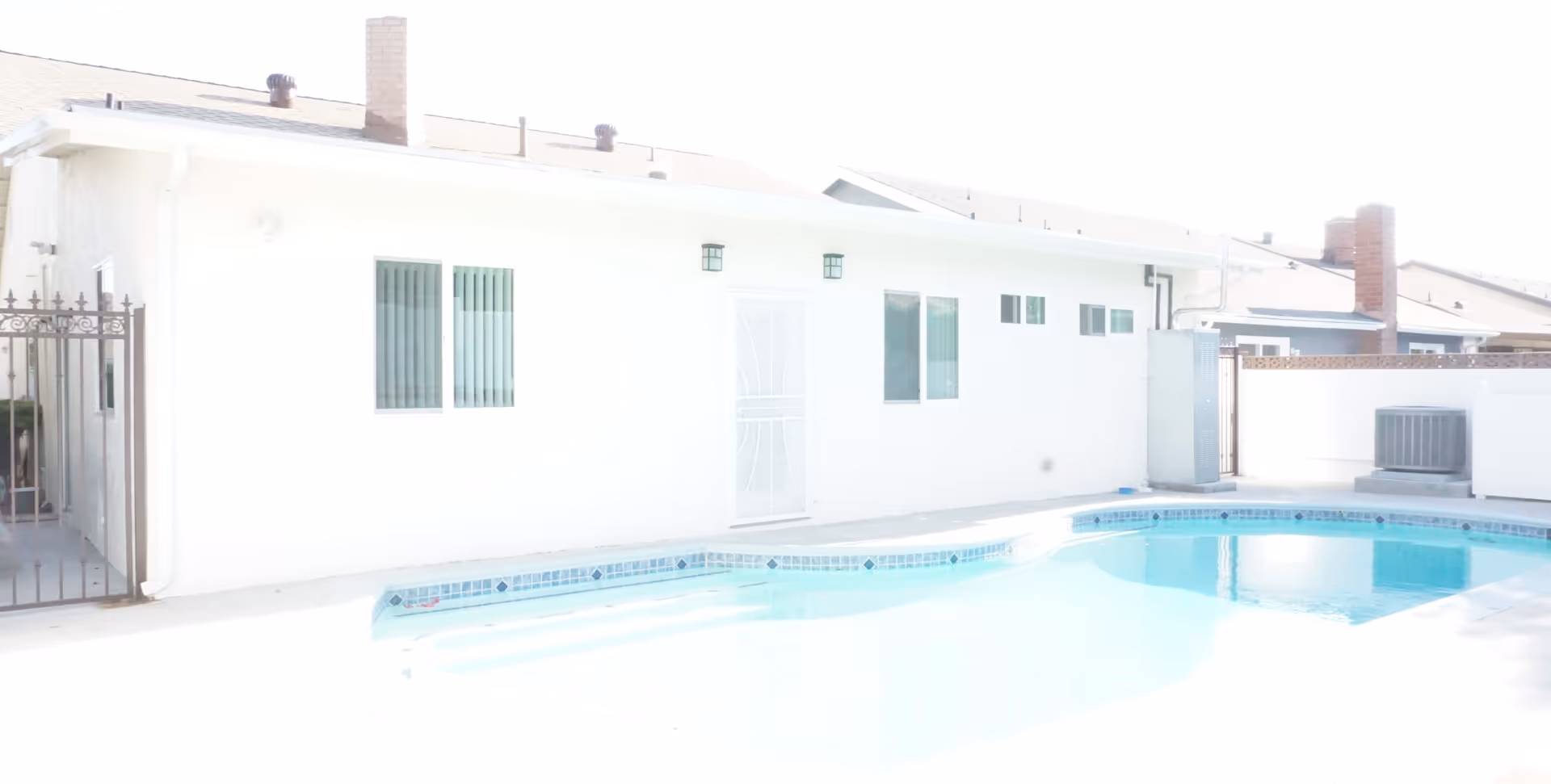 Outdoor view of a residential building with a swimming pool in the foreground. The building has white walls, several windows with vertical blinds, a white door with a decorative metal security screen, and two wall-mounted lights. There is a metal gate on the left side and an air conditioning unit on the right side near a brick chimney.