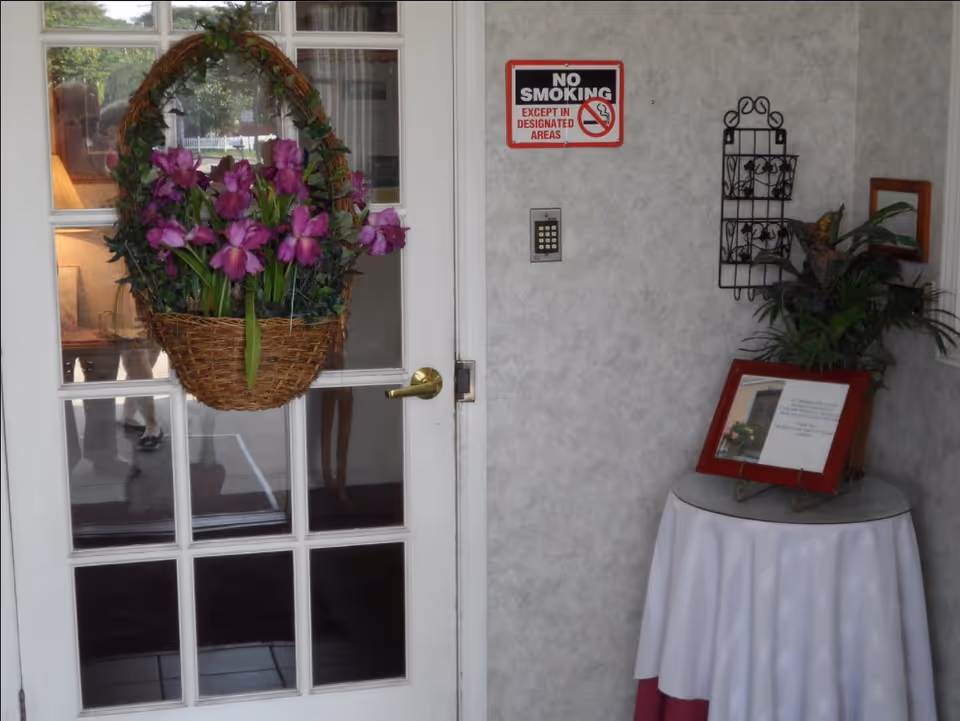 Interior view of a corner in a facility with a white door featuring glass panes and a hanging basket with purple flowers. A 'No Smoking Except in Designated Areas' sign is mounted on the wall above a keypad. A small round table covered with a white and red tablecloth holds a framed notice and a potted plant. A decorative black metal wall rack is also mounted on the wall.