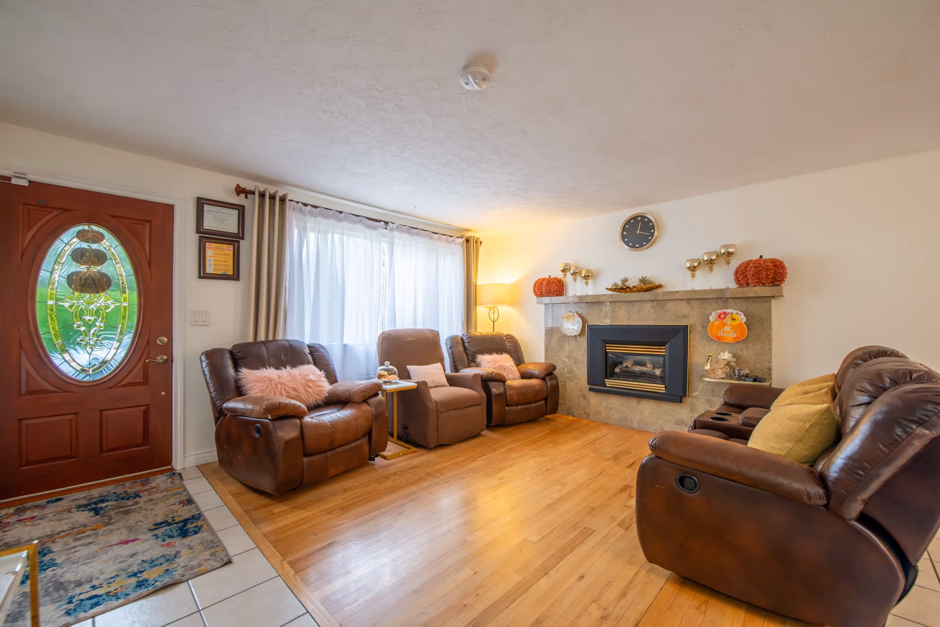 A cozy living room with four recliner chairs arranged around a fireplace. The fireplace mantel is decorated with autumn-themed items, including orange pumpkins and gold candle holders. A clock hangs above the fireplace. There is a wooden front door with an oval glass design on the left side, and a window with sheer white curtains letting in natural light. The floor is a combination of light wood and white tiles, and a colorful rug is partially visible near the door.