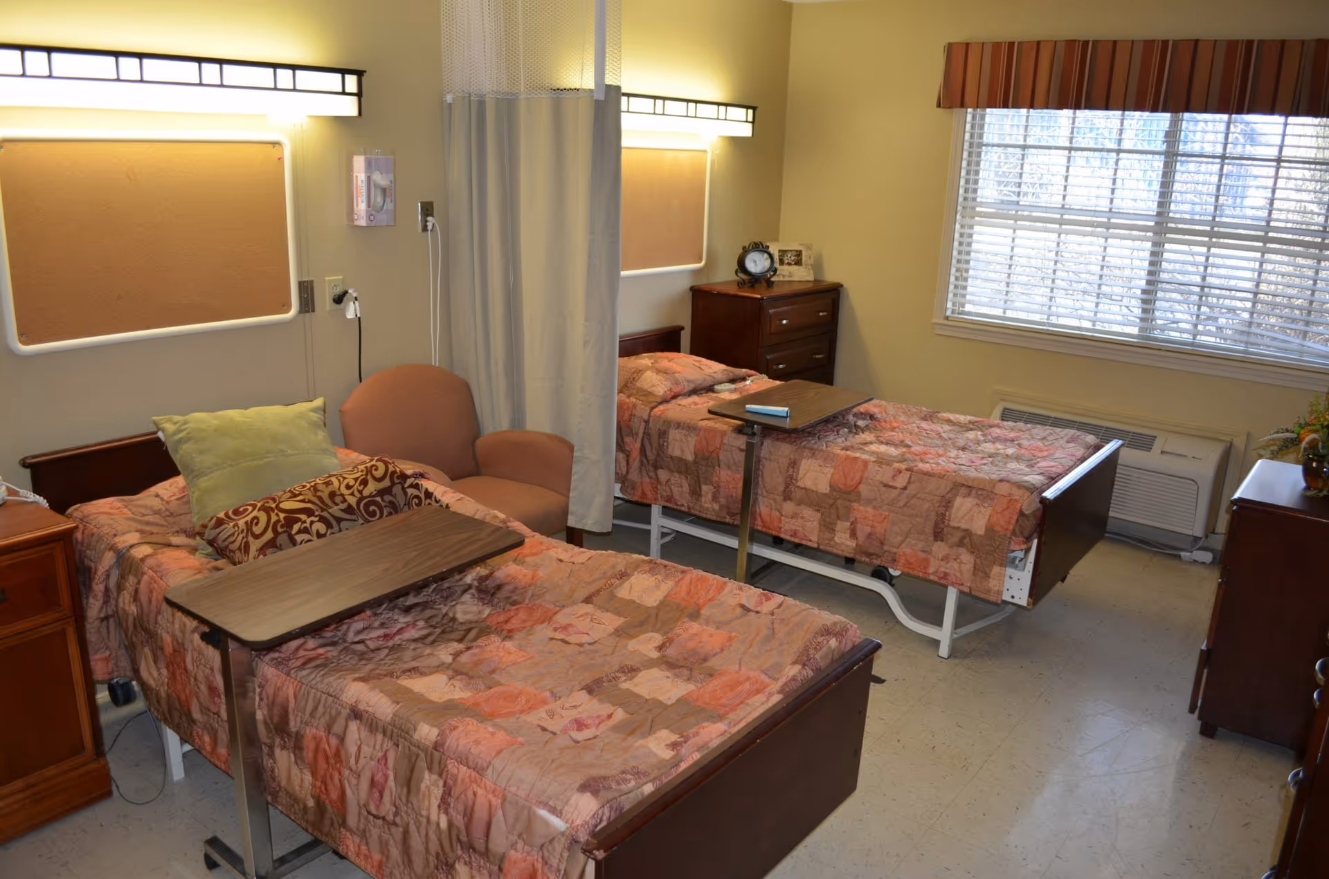 A shared senior living bedroom with two hospital-style beds, bedside furniture, a chair, and a window.