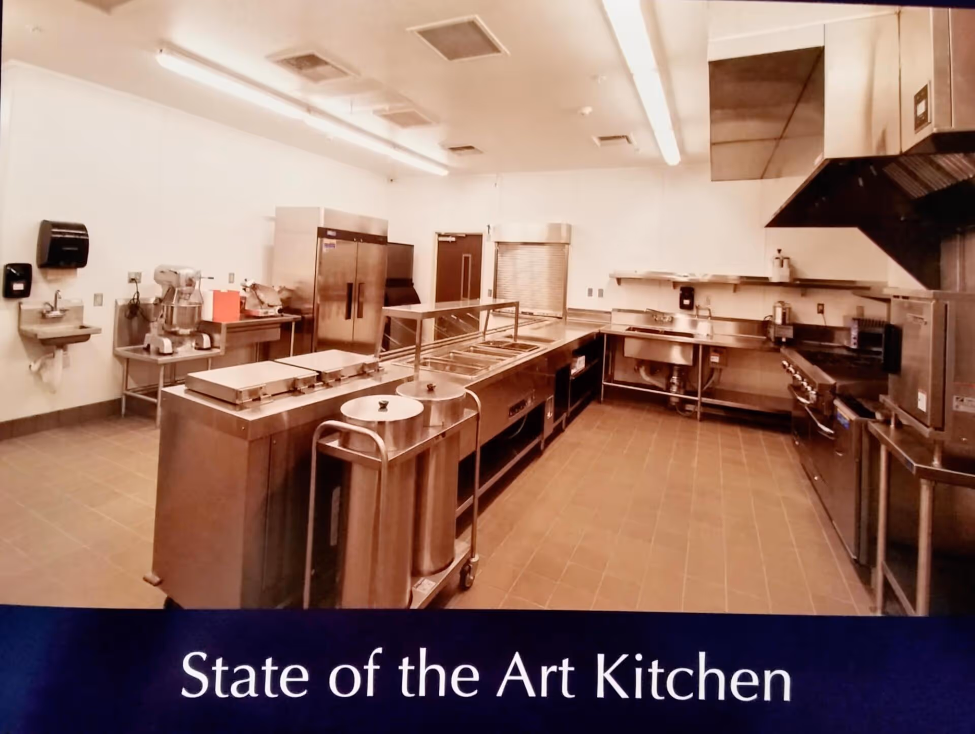A spacious, modern commercial kitchen with stainless steel appliances and workstations, including a large refrigerator, multiple sinks, cooking ranges, and food preparation areas. The kitchen is well-lit with overhead fluorescent lighting and has tiled flooring.