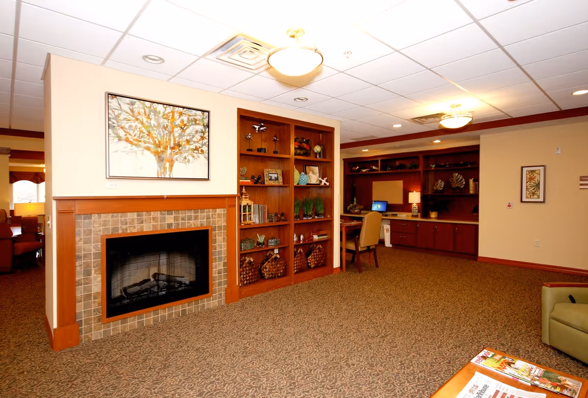 A cozy interior room featuring a fireplace with a tiled surround and a framed abstract tree painting above it. To the right of the fireplace is a wooden built-in bookshelf with various decorative items and plants. Further back, there is a desk area with a computer and chair, surrounded by additional wooden cabinetry and shelving. The room has a patterned carpet and warm lighting from ceiling fixtures.