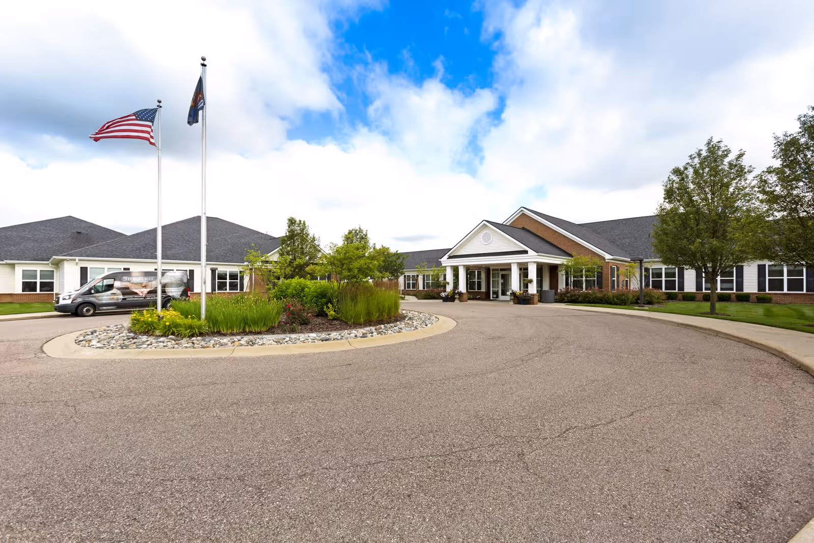 Exterior view of Olivia's Assisted Living facility showing a single-story building with a covered entrance, two flagpoles with the American flag and another flag, a circular driveway, landscaped greenery, and a van parked on the left side.