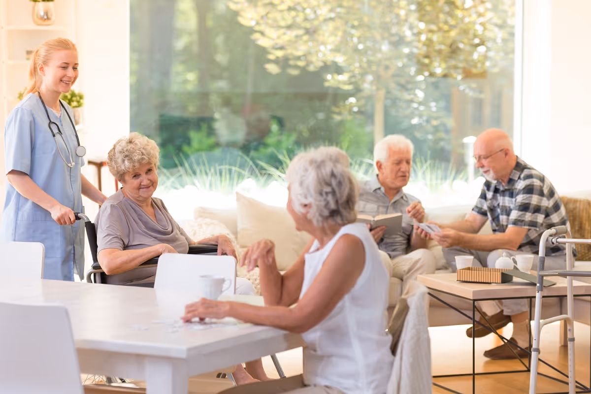 A group of elderly people socializing in a bright living room. One elderly woman is in a wheelchair being assisted by a smiling nurse in scrubs. Two elderly men are sitting on a couch playing cards, while another elderly woman is sitting at a table with a cup in front of her. The room has large windows with greenery visible outside.