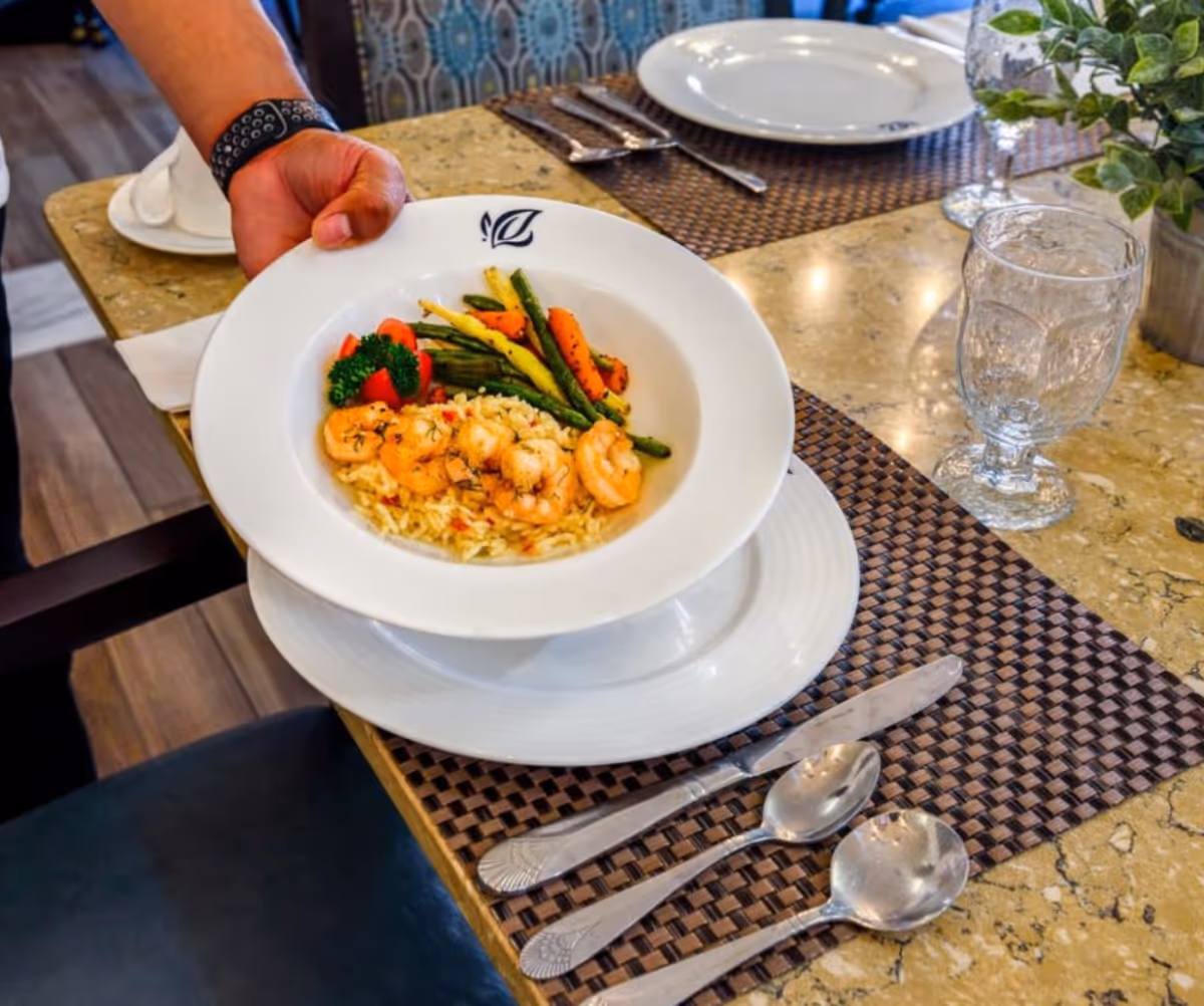 A hand holding a white bowl filled with shrimp, rice, and mixed vegetables over a dining table set with plates, silverware, a glass, and a small plant.