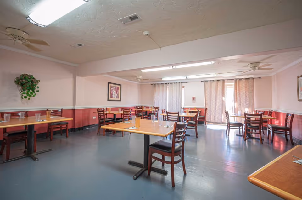 A dining room with several wooden tables and chairs arranged neatly. Each table has a glass and some utensils placed on it. The room has light pink walls with a darker pink wainscoting, a few framed pictures on the walls, and curtains covering the windows. Ceiling fans and fluorescent lights are visible on the ceiling.