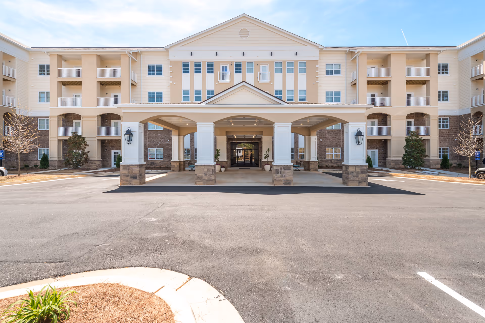 Front exterior view of a large, multi-story senior living facility with a covered entrance supported by columns. The building has balconies, multiple windows, and a paved driveway leading up to the entrance under a partly cloudy sky.