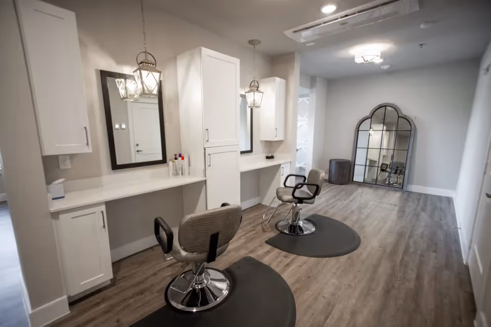 Interior view of a salon area with two styling chairs placed in front of two mirrors mounted on a white wall with cabinets. The floor is wood with protective mats under the chairs. A large decorative mirror leans against the far wall, and the space is lit by ceiling lights and hanging lantern-style fixtures.