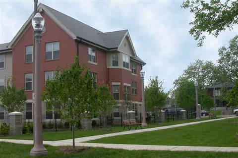 Exterior view of a three-story red brick building with multiple windows, surrounded by a green lawn, young trees, a sidewalk, benches, and street lamps under a partly cloudy sky.