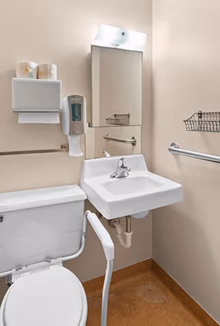 A small, accessible bathroom with a white toilet equipped with a support rail, a white wall-mounted sink with a single faucet, a mirror above the sink, a soap dispenser, a paper towel dispenser with two rolls of paper towels, a metal grab bar on the wall, and a wire basket for storage.