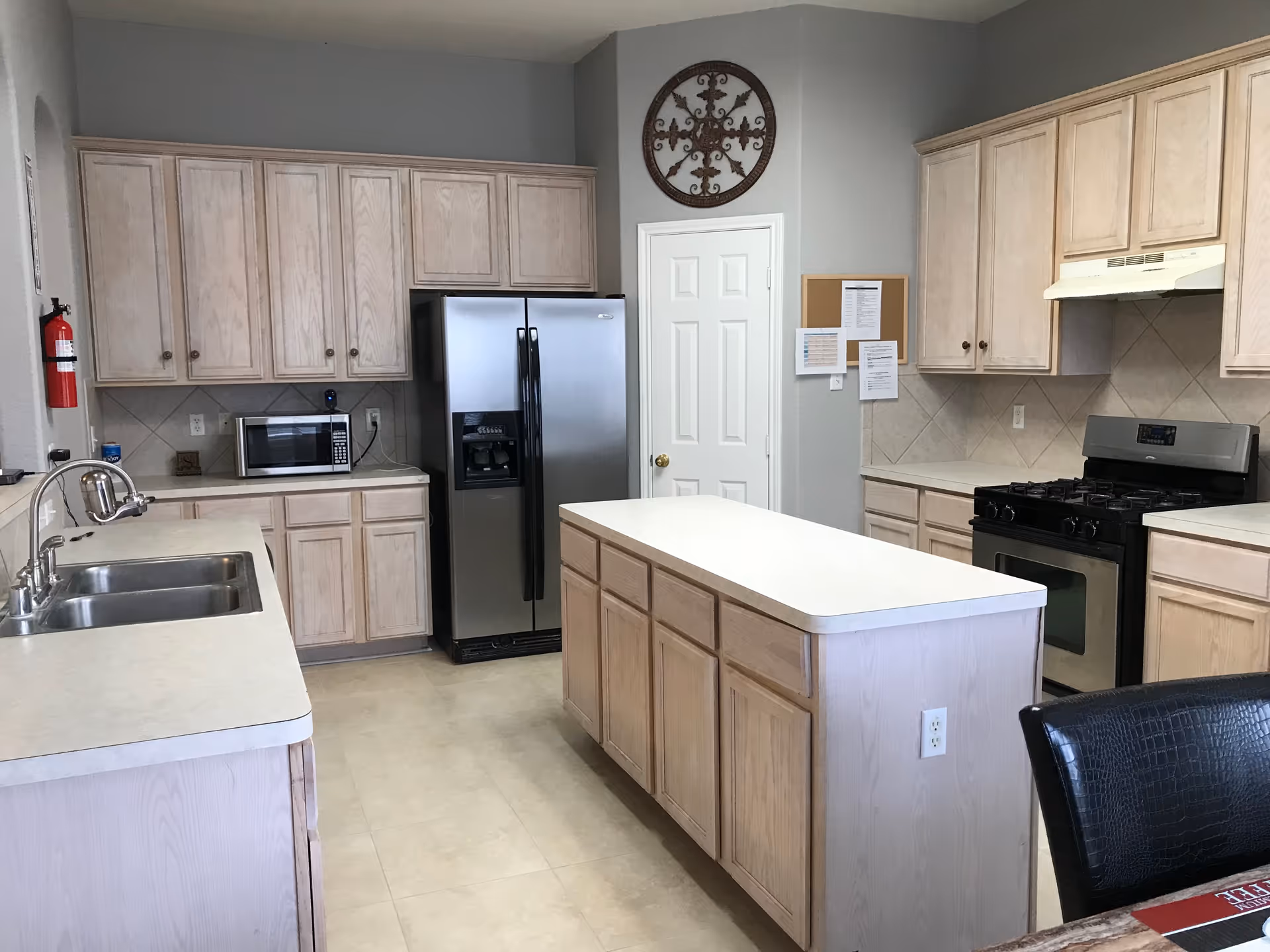 A clean kitchen with light wood cabinets, a central island, a double sink, a stainless steel refrigerator, a microwave, a gas stove with an overhead vent, and a decorative wall piece above a white door.