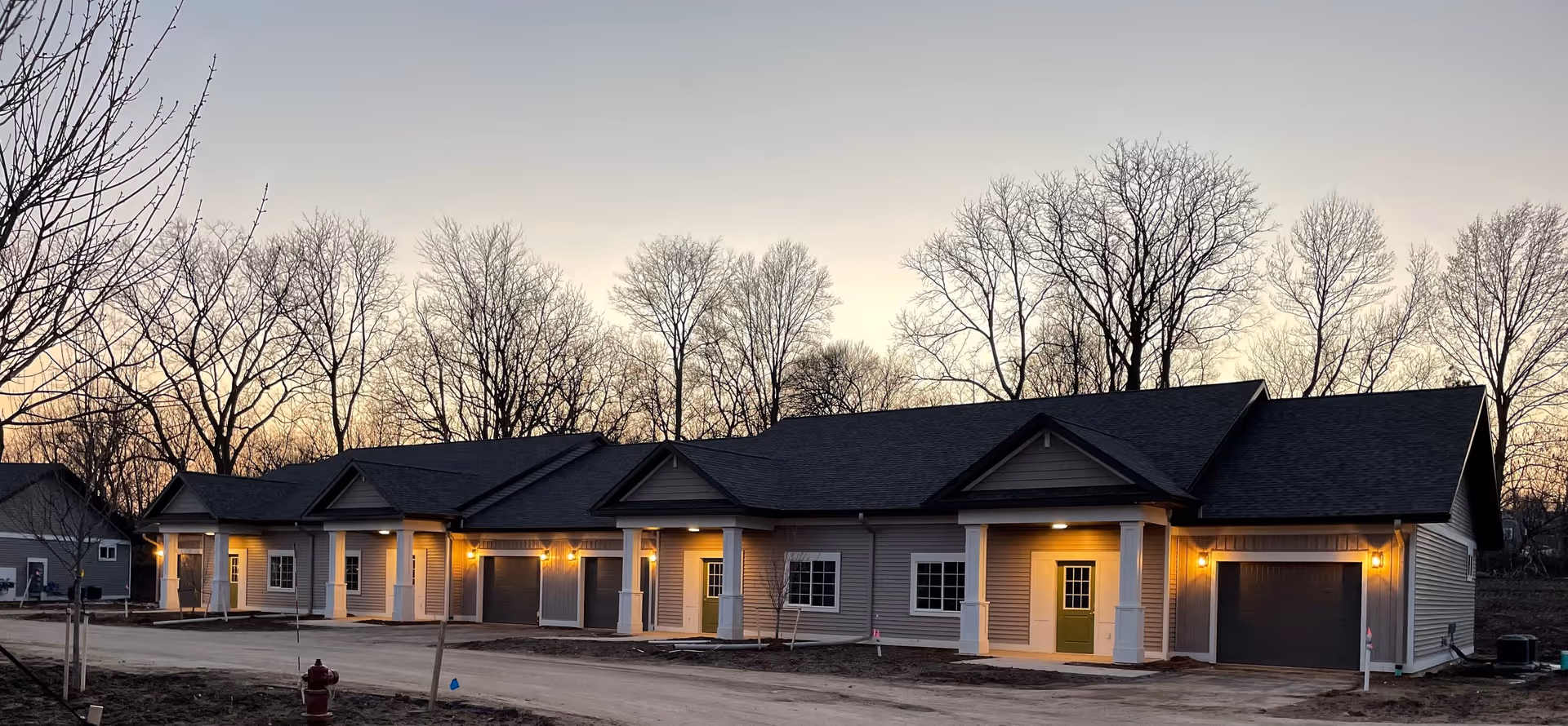 Row of single-story attached residential units with front porches and garages lit at dusk and bare trees behind.