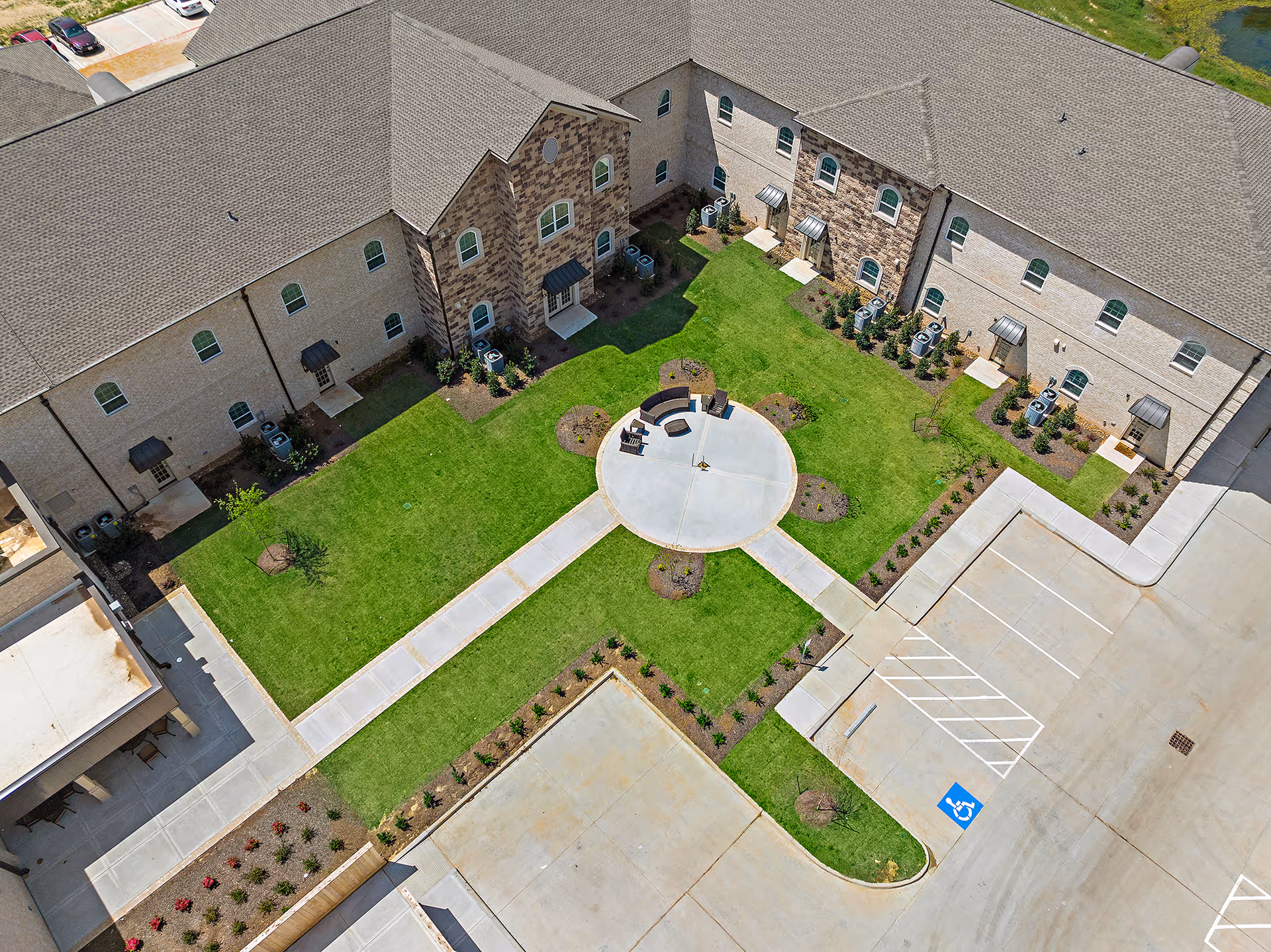 Aerial view of Worthington Manor showing a landscaped courtyard with green grass, a circular concrete seating area with benches, and surrounding pathways. The building has multiple windows and entrances with small awnings. Adjacent to the courtyard is a parking lot with marked spaces including a handicapped parking spot.
