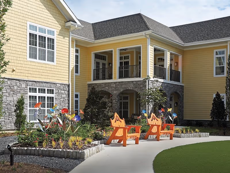 Outdoor seating area in front of a yellow and stone senior living facility building with two orange wooden chairs, colorful flower sculptures, and landscaped garden beds along a curved concrete pathway.